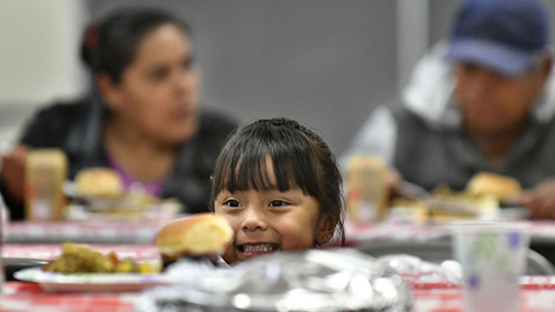 A little girl smiles broadly, her face peeking up behind her plate