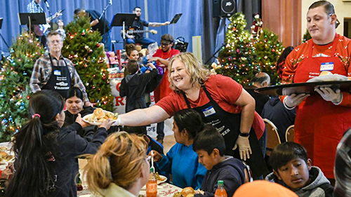 Volunteers wearing red shirts and black aprons distribute food to seated guests during the holidays