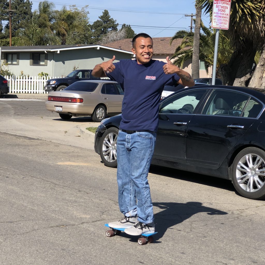 Victor holds two thumbs, smiling and riding a skateboard