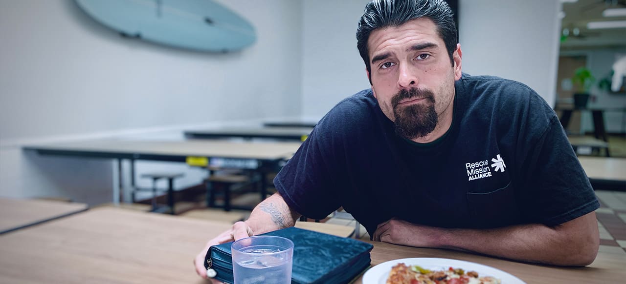 A man with a goatee sits at a table with a plate of food and a glass of water in a room with a surfboard on the wall.