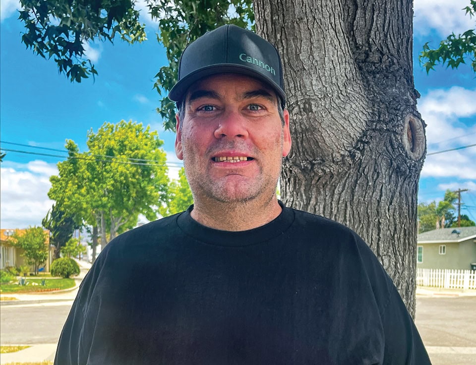Man standing outdoors near a tree with a bright blue sky in the background.