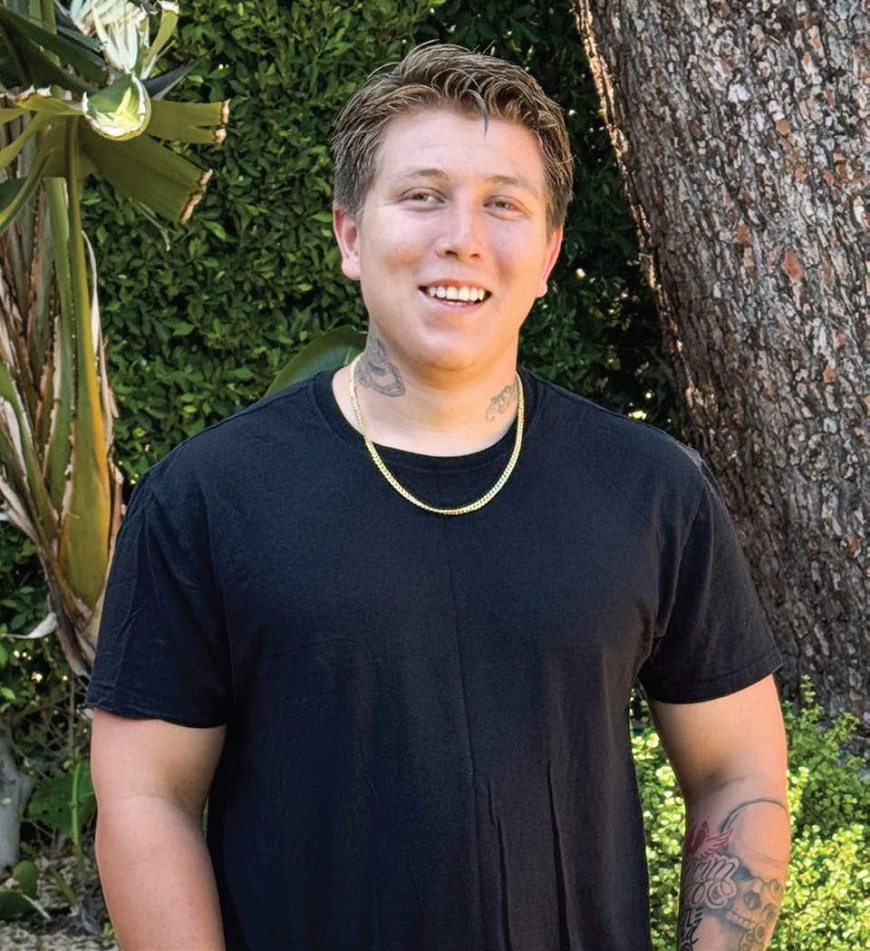 Young man smiling outdoors near a tree and lush greenery.
