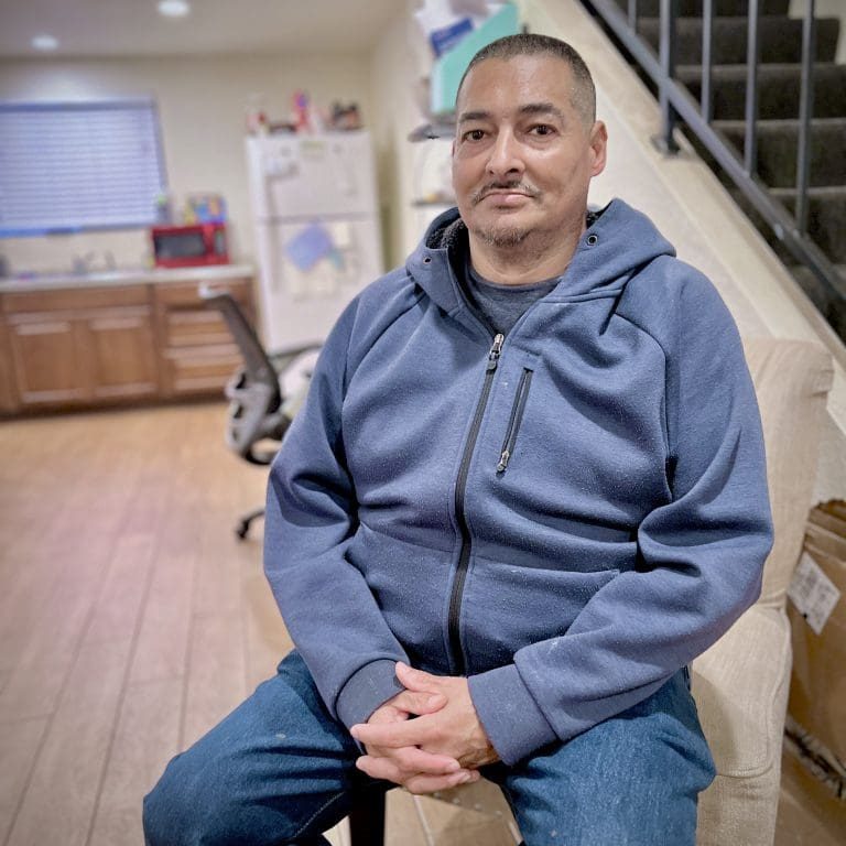 A man in a blue hoodie and jeans sits on a beige chair in a living room with wooden floors, contemplating the intersection of life’s challenges as the staircase looms in the background.