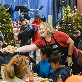 Volunteers wearing red shirts and black aprons distribute food to seated guests during the holidays
