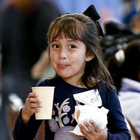 A young girl, hands full with her food and drink, smiles for a photo