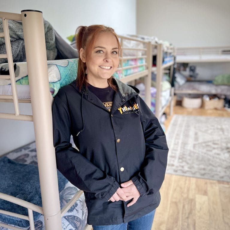 A woman stands in a room with bunk beds, exuding warmth and gratitude for Gods grace, wearing a black jacket and jeans, smiling at the camera.