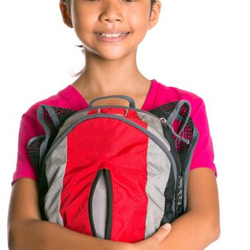 A smiling young girl with both arms wrapped around her red and grey backpack