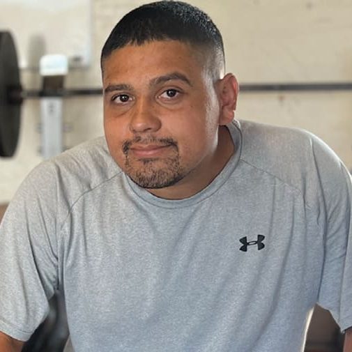 A person in a gray Under Armour shirt stands in front of gym equipment, looking towards the camera with a sense of mission and acceptance.