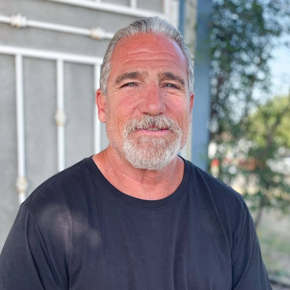 Older man with gray hair and beard smiling outdoors in front of a building.