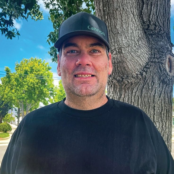 Man standing outdoors near a tree with a bright blue sky in the background.